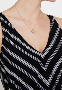 Collier en or avec pendentif circulaire, présentant un design complexe. Porté avec un vêtement à rayures noires et blanches en forme de V. Chaîne lisse et délicate.