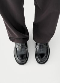 Svarte skinnloafers med glatt finish, dekorative sømmer og et vevd dekor på forsiden, kombinert med mørke dressbukser.