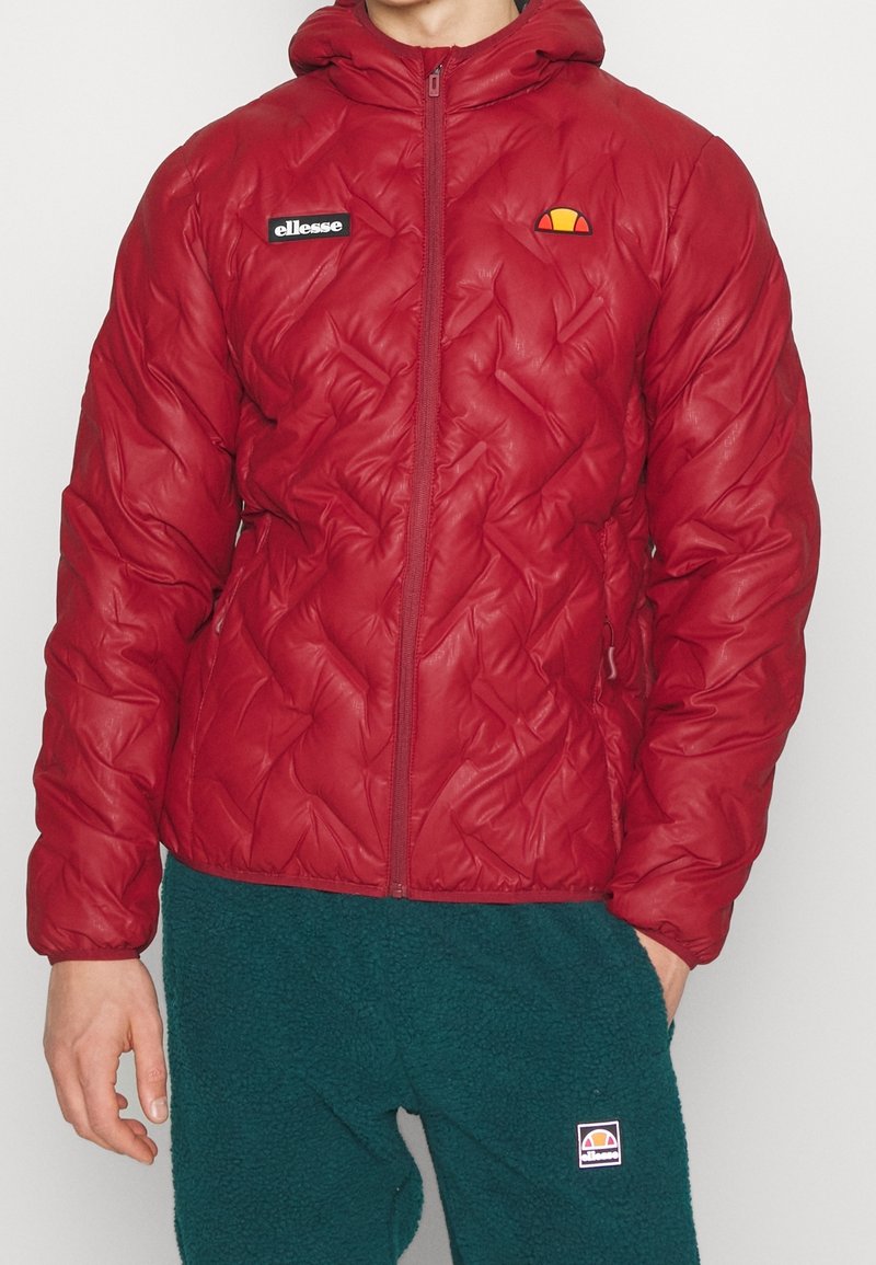Personne portant une veste matelassée rouge à capuche avec fermeture éclair et logos, associée à un pantalon en polaire vert foncé avec un logo sur la cuisse gauche.
