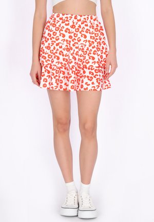 Kvinde iført en hvid og rød mininederdel med leopardprint, kombineret med hvide platformsneakers og ankelstrømper, stående mod en hvid baggrund.