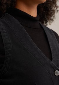 Černá kordurová vesta s výstřihem do V a ramenními popruhy, zdobená kovovým knoflíkem. Nositá přes černý rolák.