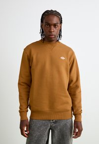 Felpa marrone con collo e polsini a coste, con un piccolo logo bianco sul lato sinistro del petto. Indossata con jeans grigi sbiaditi.