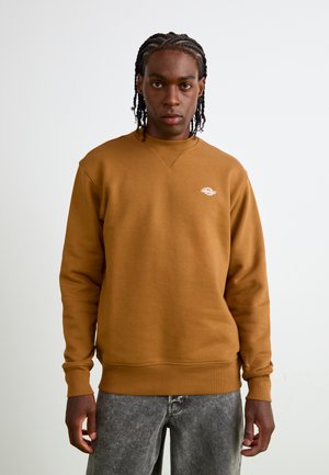 Sweatshirt maro cu guler și mansete ribbate, având un mic logo alb pe partea stângă a pieptului. Purtați-l cu blugi gri decolorati.