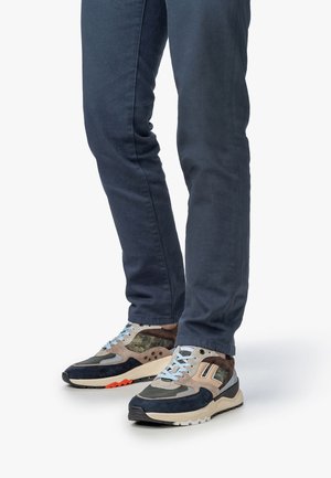 Hombre con pantalones azul oscuro y zapatillas multicolores con cordones beige, navy, camuflaje y azul claro, de pie sobre un fondo blanco.