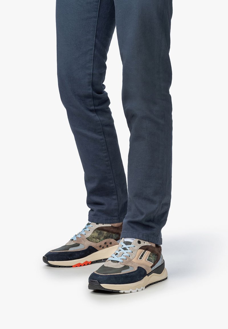 Uomo con pantaloni blu scuro e scarpe da ginnastica multicolori con lacci beige, blu navy, mimetici e azzurri che si trova su uno sfondo bianco.