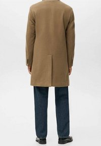 Manteau en laine marron au design droit, avec une fente unique à l'arrière et des boutons noirs en accent, associé à un jean bleu foncé et des chaussures noires.