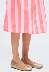 Πόδια με ροζ και κοραλλί ριγέ φούστα σε συνδυασμό με μεταλλικά χρυσά μπαλαρίνια Mary Jane σε λευκό φόντο.