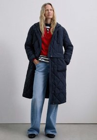 Quilted navy puffer coat with a hood, front pockets, and snap closure, layered over a red sweater and striped shirt, paired with light blue jeans.