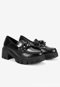 Mocasines de cuero negro con suela gruesa, que presentan un acento decorativo de lazo en la parte superior y una textura suave y brillante.