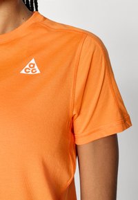 Πορτοκαλί κοντομάνικη μπλούζα από βαμβάκι, με τρίγωνο λευκό λογότυπο στην πάνω αριστερή πλευρά και ραφή flatlock κατά μήκος των ενώσεων.