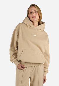 Hanorac oversized bej, realizat dintr-un material moale, având o glugă mare, buzunar frontal și un logo imprimat. Mansete și tiv cu nervuri.