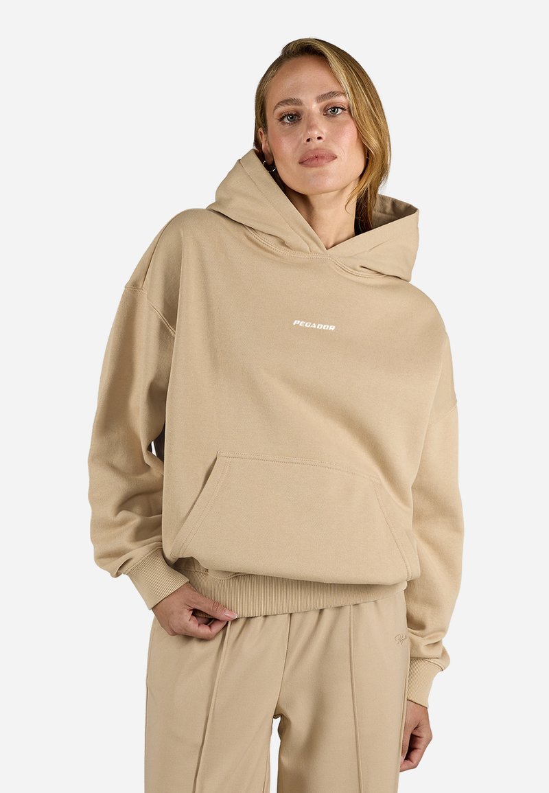 Hanorac oversized bej, realizat dintr-un material moale, având o glugă mare, buzunar frontal și un logo imprimat. Mansete și tiv cu nervuri.