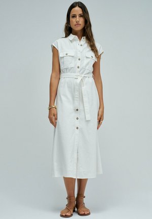 Frau steht und trägt ein ärmelloses weißes midi-Knopfkleid mit gebundenem Taillengürtel und braunen flachen Riemchensandalen.