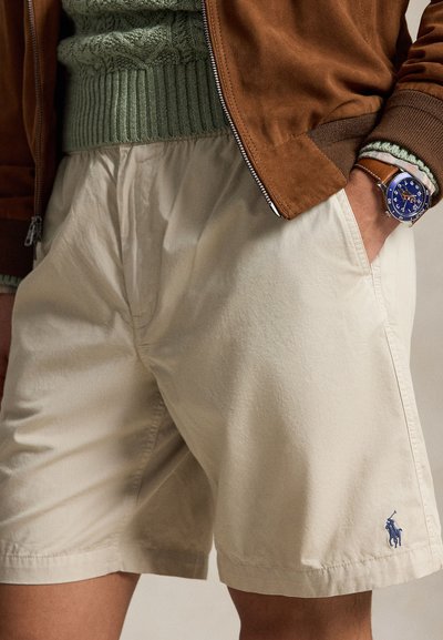 Bež kratke hlače z modrim logotipom polo, nošene z zelenim puloverjem in rjavo jakno; roka osebe v žepu kaže uro z modrim številčnico in rjavim paščkom.