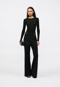 Mulher em pé, vestindo uma blusa preta ajustada de mangas longas, calças largas pretas de cintura alta com detalhes em botão, segurando uma pequena bolsa preta.