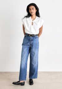 Witte top met een keyhole halslijn, brede schouders, gecombineerd met hoge taille, lichtblauwe, wijde spijkerbroek en zwarte loafers.