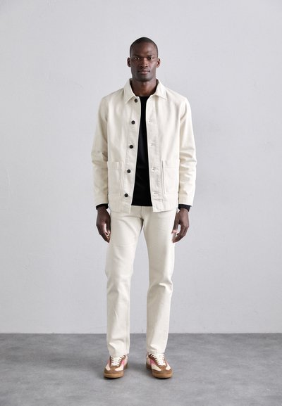 Off-white canvas jacket with black buttons and pockets, paired with matching trousers and multicolored sneakers, worn by a standing man.