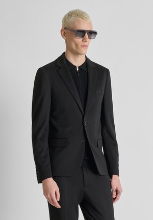 Man met kort blond haar, grote zwarte zonnebril, zwarte blazer en zwart vest met rits, tegen een effen grijze achtergrond.