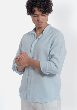 Giovane uomo con capelli ricci in camicia azzurro chiaro con bottoni, guarda verso il basso, con le mani intrecciate davanti a uno sfondo bianco uniforme.