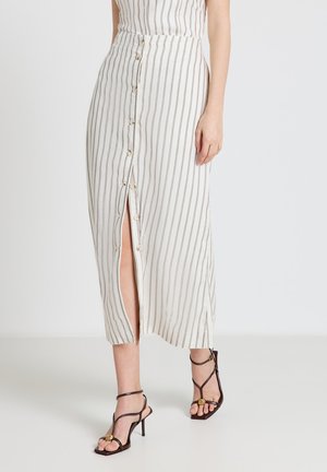 Frau trägt einen weißen Midirock mit dünnen vertikalen Streifen, Vorderknöpfen und einem Schlitz, kombiniert mit schwarzen, Riemchen-High-Heel-Sandalen.