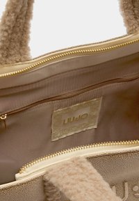 Beige textured handbag with fluffy handles, gold zipper, and inner zip pocket. LIU JO logo inside visible on a cream background.