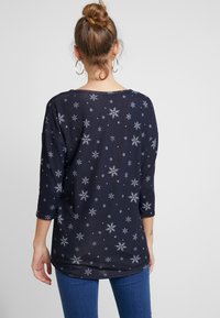 Haut bleu marine à manches longues avec des motifs de flocons de neige argentés, col rond, coupe décontractée, porté avec un jean bleu.