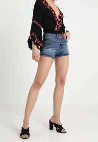 Svart blus med röd broderi och klockärmar, i kombination med ljusblå denimshorts. Modellen bär svarta klacksandaler.