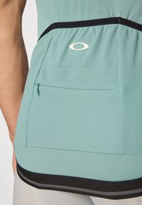 Lys blågrønn trenings tanktop med strukturert stoff, som har en svart aksentkant og en liten glidelåslomme på baksiden.