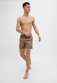 Barna úszónadrág, rugalmas derékrésszel és húzózsinórral, a elején egy kis fehér logóval. A modellen mezítláb van.