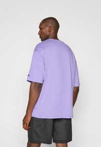 Tricou din bumbac purpuriu, cu mâneci scurte, guler rotund și o croială lejeră. Purtați cu pantaloni scurți din plasă negri, evidențiind un design simplu.