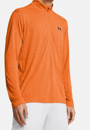 Mężczyzna w jaskrawo pomarańczowej sportowej koszulce z długim rękawem, z subtelnym geometrycznym wzorem i suwakiem do ćwierci długości, w połączeniu z białymi spodniami.