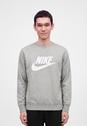 Jonge man die een grijze Nike-sweatshirt met wit logo draagt, staand tegen een effen lichte achtergrond, met een lichte glimlach naar voren gericht.