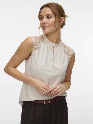 Naine kannab varrukateta beežikat topi pitsdetailidega õlgadel ja musti pükse pruunide vööga, vaatab vasakule.