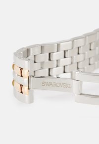 Bracciale per orologio in metallo argento e dorato con anelli intrecciati, dotato di una chiusura incisa con la scritta "SWAROVSKI". Texture liscia e finitura lucida.