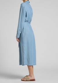 Henkilö, joka wearoksiaalilla vaaleansinisessä pitkässä farkkuhaalarissa, jossa on sivuvaie, beigeissä sandaaleissa, esitettynä sivuprofiilissa yksinkertaista taustaa vasten.