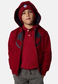 Sudadera roja con capucha y forro azul marino, bolsillos delanteros y logo; combinada con una camiseta polo roja. El niño lleva un gorro de lana rojo a juego.
