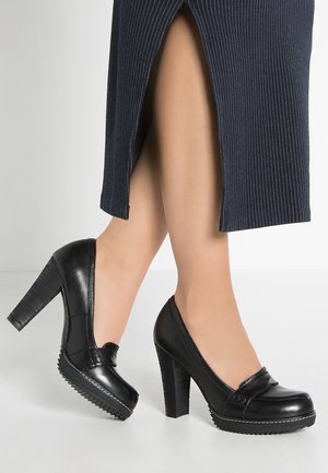 Femme portant des mocassins noirs en cuir à talons hauts épais et semelles rainurées, assortis à une jupe bleu marine à rayures fines avec une fente à l'avant.