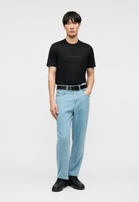 Άνδρας που φοράει μαύρο κοντομάνικο πουκάμισο, ανοιχτόχρωμο μπλε τζιν με μαύρη ζώνη και μαύρα παπούτσια, στέκεται με το ένα χέρι στην τσέπη μπροστά σε λευκό φόντο.