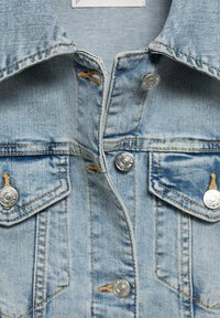 Lysblå denimjakke med sølvfarvede metaltrykknapper, to brystlommer og en spids krave. Distressed teksturer synlige på stoffet.