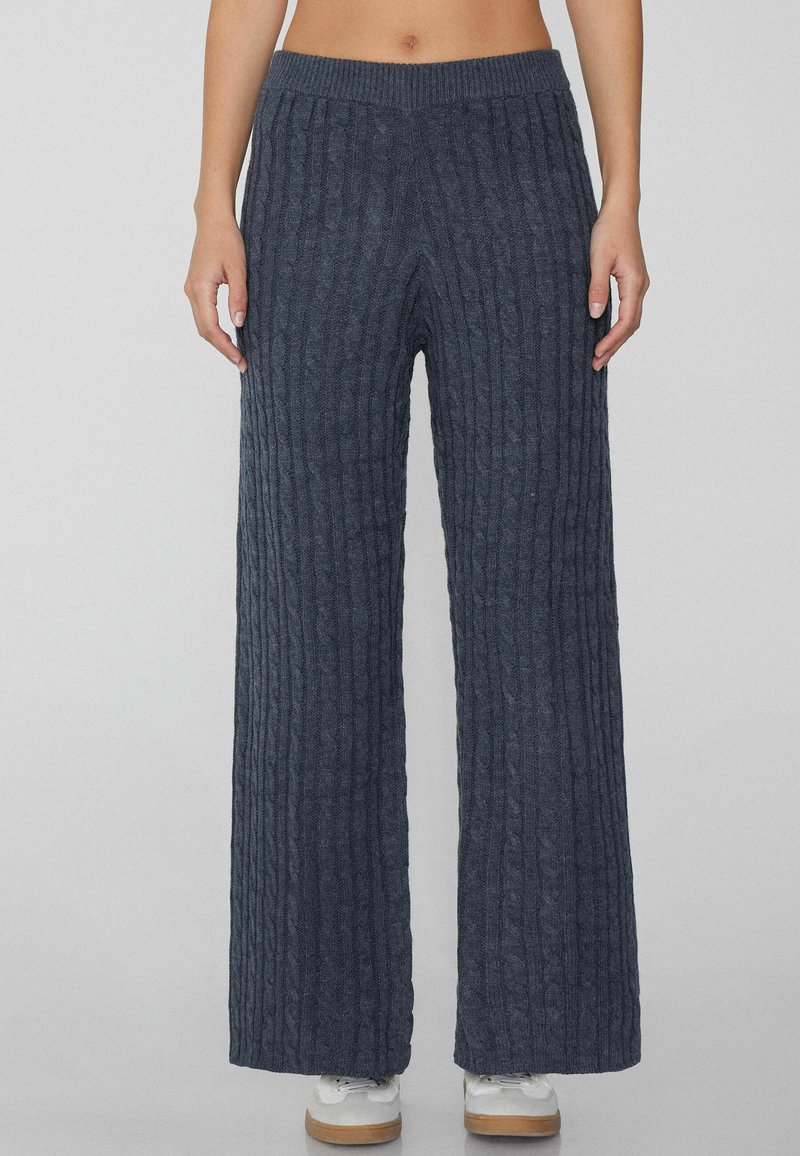 Pantalones de punto grueso de pierna ancha en gris oscuro con textura de canalé vertical y cinturilla elástica. Combinados con zapatos blancos.