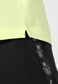 Světle žlutá texturovaná košile s bočními rozparky, kombinovaná s černými šortkami s opakujícím se bílým logem krokodýla po celém pásu.