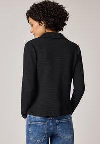 Femme aux cheveux bouclés vue de dos, portant un haut noir texturé à manches longues et un jean bleu en denim.