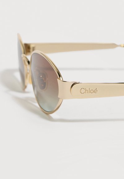 Γυαλιά ηλίου με χρυσή κορνίζα, φακούς με βαθμιαία σκουρόχρωμη απόχρωση και χαραγμένο το όνομα «Chloé» στα πλατιά πλαϊνά μπράτσα, τοποθετημένα σε λευκή επιφάνεια.