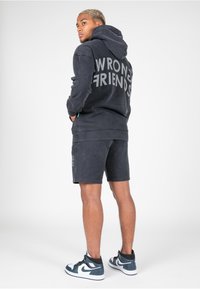 Sudadera negra con capucha y pantalones cortos con el texto "WRONG FRIENDS", fabricados en algodón, que cuenta con capucha y bolsillos. Llevado con zapatillas altas.