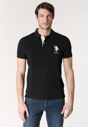 Uomo che indossa una polo nera USPA e jeans blu, in piedi contro uno sfondo chiaro e uniforme, rivolto verso la fotocamera.