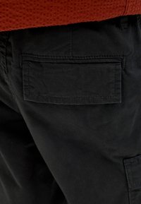 Gros plan sur une poche de pantalon noire avec un détail de rabat, portée avec un haut orange texturé.