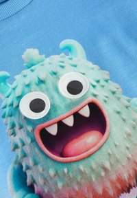 Modrá mikina s půvabnou zelenou chlupatou příšerou s velkýma plastovýma očima, ostrými zuby a usměvavou pusou, která dodává hravý detail designu.