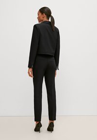 Schwarze, zugeschnittene Jacke mit einem Tailored-Design, kombiniert mit schmal geschnittenen schwarzen Hosen und High Heels. Glatter Stoff, klare Linien, minimale Details.