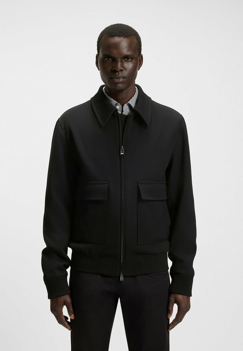 Veste bomber noire avec fermeture éclair à l'avant, grandes poches avant, poignets et ourlet côtelés, fabriquée dans un tissu lisse et structuré.