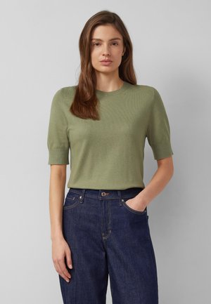 Vrouw met lang bruin haar, gekleed in een olijfgroen gebreid shirt met korte mouwen en een high-waisted donkerblauwe spijkerbroek, staand tegen een effen achtergrond.